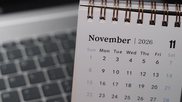 Close-up of a desk calendar on a laptop keyboard as a hand flips the page from October 2026 to November 2026. Camera stays static, showing a clear monthly time transition and structured planning conc