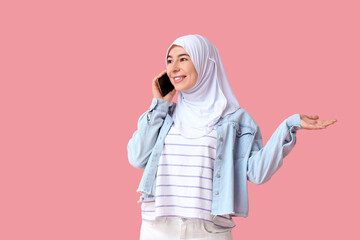 Fototapeta premium Beautiful young Muslim woman in hijab talking by mobile phone on pink background