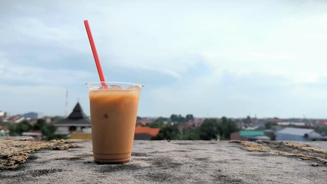 A plastic cup of iced coffee with a red straw placed on a concrete rooftop, overlooking a quiet urban neighborhood under a bright blue sky.