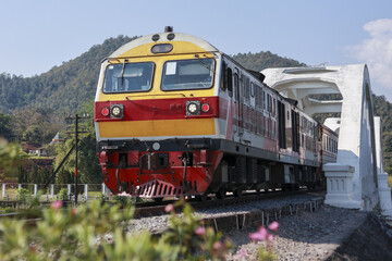 Obraz premium Vibrant yellow and red train on scenic journey, crossing concrete railway bridge. This powerful locomotive travels through lush green mountain landscape of rural Thailand
