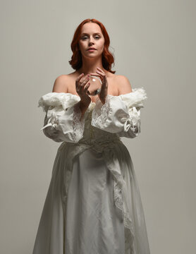 close up portrait beautiful redhead female model wearing historical white fantasy wedding dress, fairytale  ball gown, holding crystal seers orb. overhead studio lighting on white background