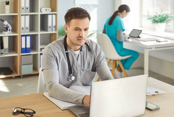 Young confident male doctor wearing uniform working at the desk with laptop on workplace in medical...