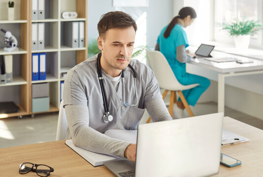 Young confident male doctor wearing uniform working at the desk with laptop on workplace in medical clinic or hospital with female nurse colleague. Medicine and health care concept.