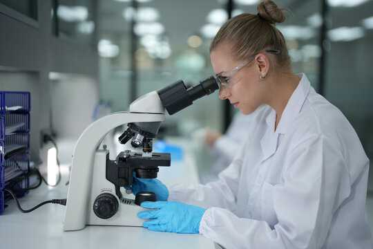 Side view of Caucasian female scientist looking through microscope in modern lab. Young researcher in safety glasses and gloves performing medical analysis.