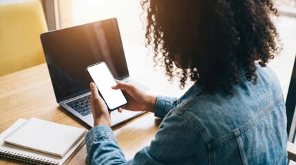 Young Black woman wearing denim jacket scrolling smartphone with blank white screen mockup beside open laptop and notebooks.