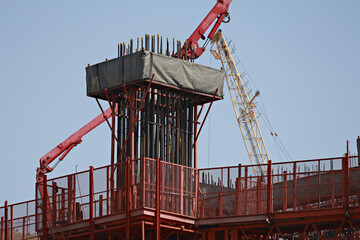 Construction site with red crane and scaffolding is shown, featuring concrete column being reinforced with steel bars. scene captures industrial process under clear blue sky