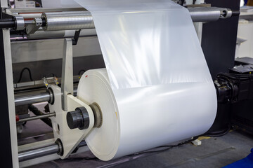 Industrial machine winding large spool of white plastic film on roller in manufacturing facility with metal components and precision engineering. Packaging material. Packaging release