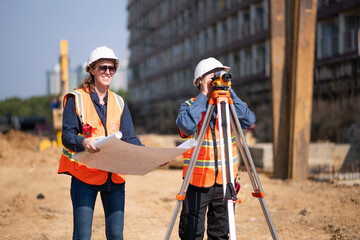 Construction surveyor or engineer working construction site blueprint and theodolite mounted tripod measuring angles and distances with heavy machinery excavator constructed hi-way road background.