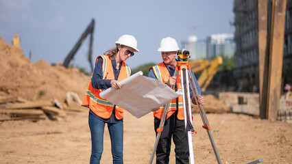 Construction surveyor or engineer working construction site blueprint and theodolite mounted tripod measuring angles and distances with heavy machinery excavator constructed hi-way road background.