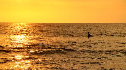 Fototapeta premium A serene summer landscape at sunset featuring the orange silhouette of a man walking along the calm ocean beach as waves reflect the golden horizon and clouds over the sea