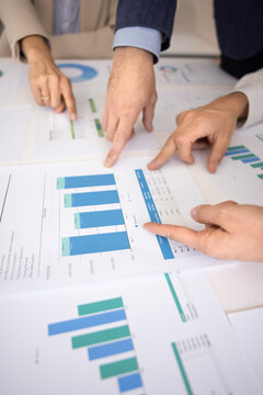 Close-up of hands pointing at diverse bars and figures on printed financial charts laid out on conference table, collaborate, analyze data trends, discussing key performance indicators during meeting