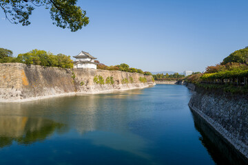 A traditional Japanese castle tower stands above impressive stone ramparts, overlooking a wide, calm moat surrounded by lush greenery under a clear blue sky in Osaka. The tranquil water reflects the