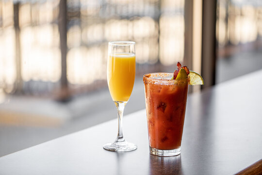 A champagne mimosa with orange juice and a bloody mary with olives in a bright window bar seat at a modern breakfast cafe 