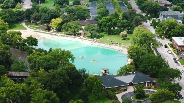 Drone footage shows large outdoor swimming pool surrounded by trees, walking paths, and park landscaping in city environment. Recreational area for leisure, summer activity