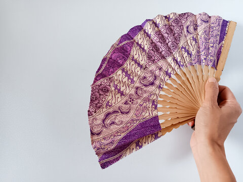 Close up of woman hand holding a purple and beige Indonesian traditional batik handheld folding fan or kipas tangan with a bamboo frame isolated on white background.