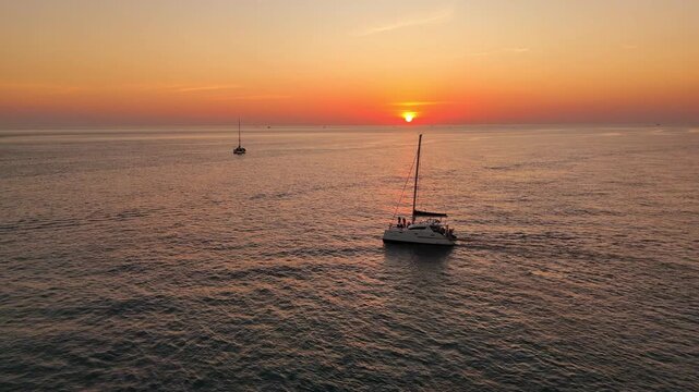 Cinematic aerial view of a sailing catamaran yacht with raised sails cruising the Andaman Sea near Phuket, Thailand. Tourists enjoy a vivid orange tropical sunset over glowing ocean waters