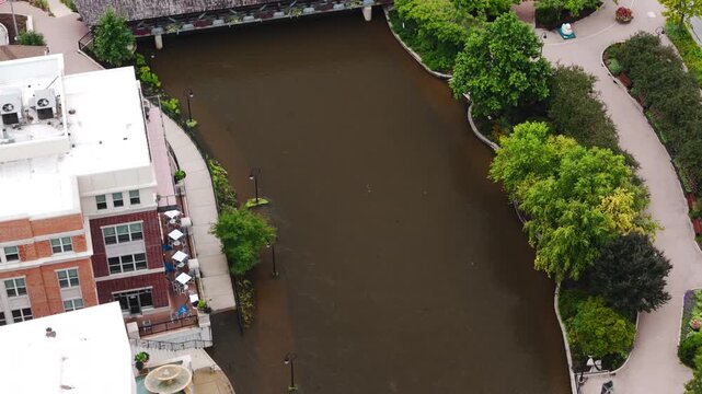 Aerial view of a swollen river flowing through a city after heavy rainfall. High water levels fill the river channel near buildings and walkways, showing flooding conditions