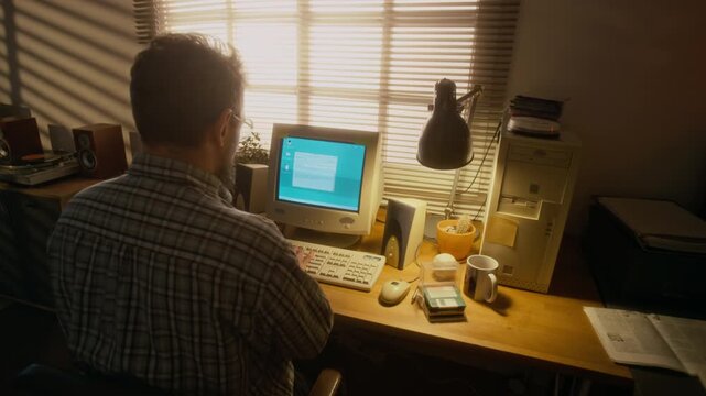 Rear shot of mature man in glasses sitting with back to camera, writing email on old-fashioned computer in cozy home study, sunlight streaming through window blinds with shadows on walls, retro vibe