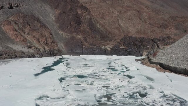 Dynamic aerial of heavily cracked white ice surface on frozen Zanskar River squeezed between towering rocky canyon walls in Ladakh Himalayas.
