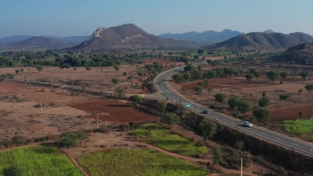 Overhead aerial tracks car moving along winding highway through arid Rajasthan landscape with scattered trees and granite hill formations near Bhangarh.