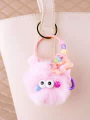 Close-up of a soft, fluffy plush pink  keychain in the shape of a monster with eyes on a white bag.