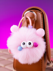 Close-up of a soft, fluffy plush pink  keychain in the shape of a monster with eyes on a brown bag.