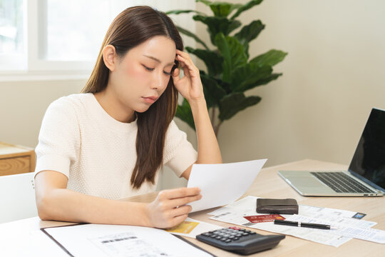 Debt and financial concept, stressed asian young woman have a headache while female calculate expenses, income and expenses, looking bills of credit card for payment or payday on table at home office.