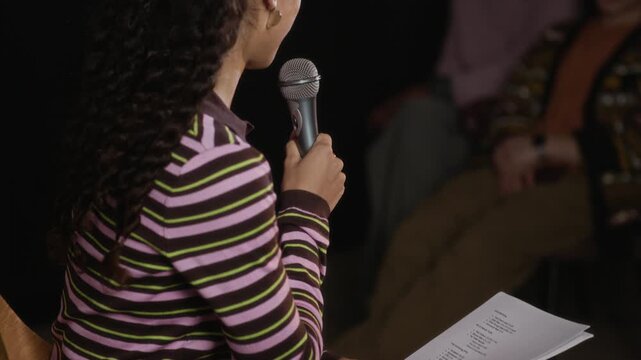 Tilt up of young Biracial woman in striped long sleeve holding microphone and reading notes from paper while speaking during public talk or event with engaged audience