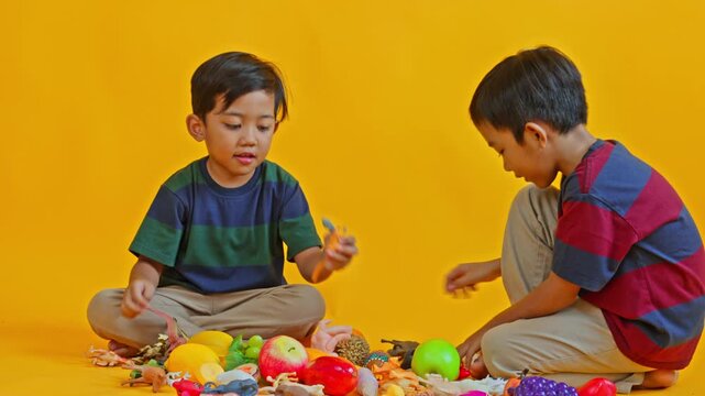 Happy young siblings sitting and playing with toy dinosaurs and plastic fruits on yellow background

