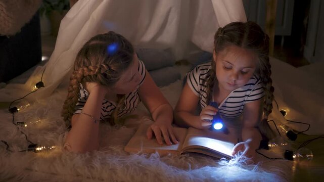 Sisters reading by flashlight inside fort, sister and girl child exploring open book on blanket rug, braided hair, cozy tent with string light glow, night indoor play, focused face, shared story