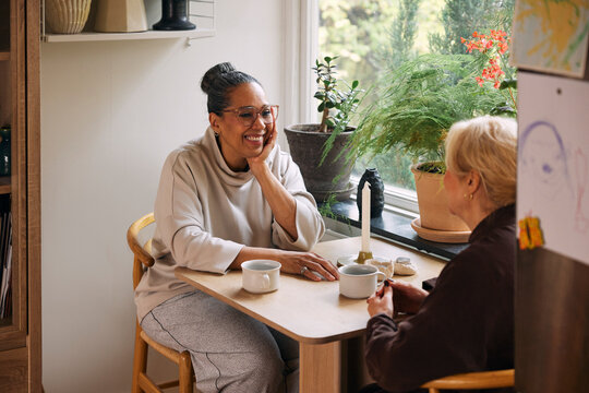 Smiling mature woman leaning on elbow while talking with female friend at table in home