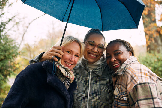 Portrait of smiling multiracial female friends standing side by side under umbrella in rain
