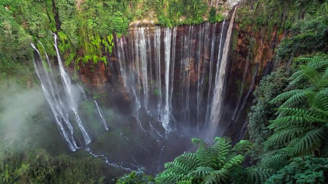 High angle drone video capturing Tumpak Sewu Waterfall in Indonesia with countless water streams flowing over steep canyon walls creating heavy mist, lush greenery and a powerful tropical landscape.