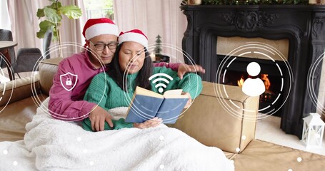 Reading couple in Santa hats cuddling at home on sofa, holding blue book and showing icons