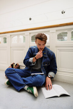 Focused young male student studying while sitting cross-legged on ground in corridor at college