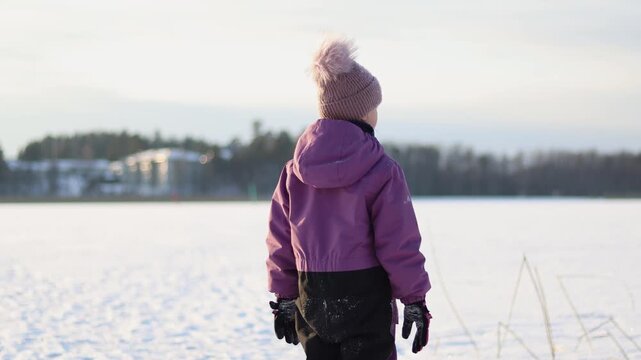 4K video of a child standing on a frozen lake in winter, looking into the distance. Snow-covered surroundings and cold light create a calm, reflective atmosphere that symbolizes curiosity, imagination