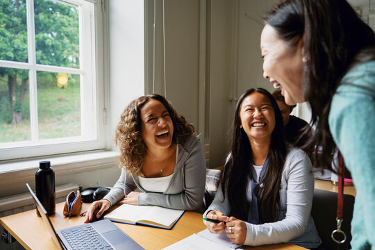 Female students laughing with teacher while sitting in classroom at college