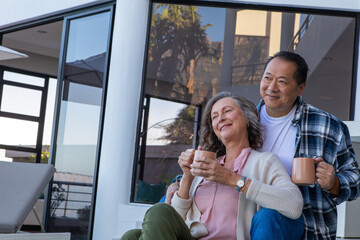 Diverse senior couple relaxing on patio balcony holding mugs near glass window, copy space