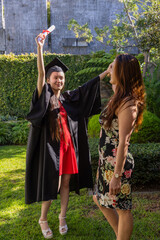 Asian mother and daughter posing in backyard in black cap gown with watch, holding diploma scroll