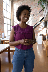 African american woman smiling, standing in office wearing purple top, jeans with pc monitor