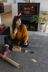 Naklejka premium Woman in her thirties wrapping gifts using kraft paper sitting on rug by fireplace
