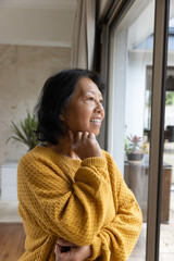 Senior Asian American woman gazing outside through sliding door inside home near potted houseplant