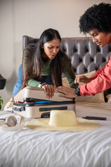 Diverse couple leaning over white bed pressing down on suitcase lid beside straw sun hat