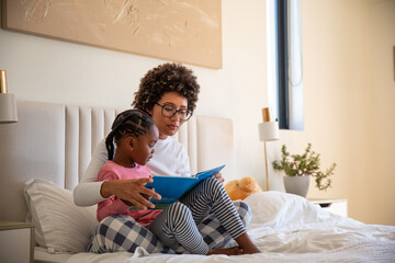 African american mother with daughter sitting on bed in pajamas reading blue storybook