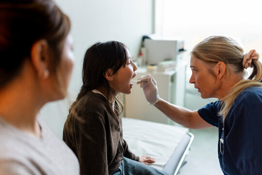 Senior female pediatrician examining girl's mouth with tongue depressor in clinic