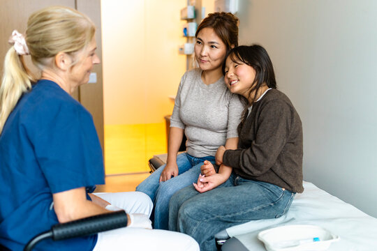 Smiling girl leaning on mother's shoulder while talking with female doctor in hospital