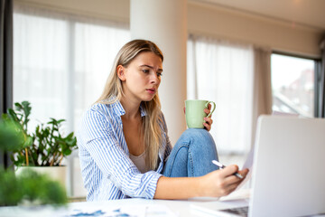 Concerned Young Woman Holding Coffee Mug and Reviewing Documents at Home Desk. Stressed Female...