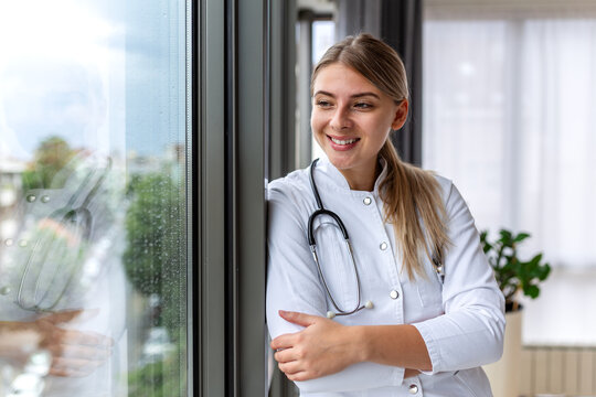 Smiling female doctor looking out window. Healthcare and medicine concept. Professional medical worker with stethoscope leaning on window. Young attractive woman practitioner in modern clinic.