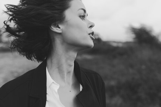 Profile of a woman in motion, wind tousling hair, wearing a blazer and white shirt, outdoor setting with a cinematic black and white mood.
