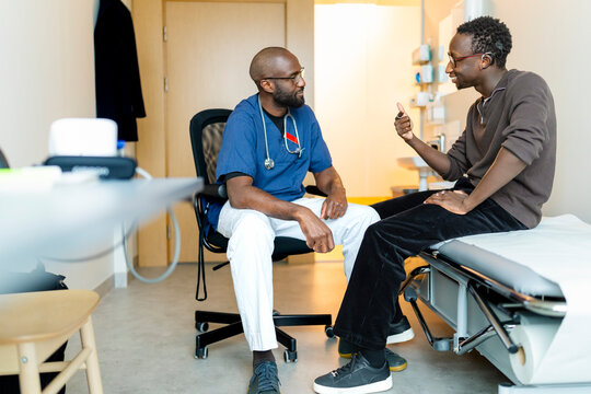 Full length of young man communicating problems through sign language to doctor while sitting on examination table in cl